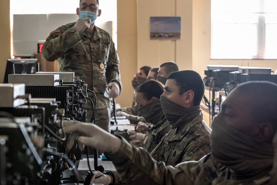 Seven service members wearing face masks sit in a row and operate radios as an instructor looks on.