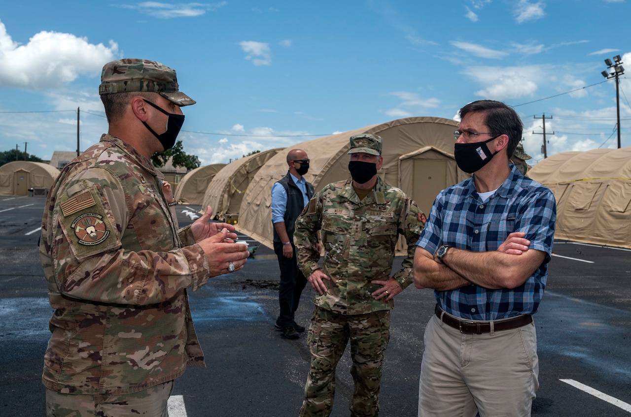 An airman and a civilian talk in a tent city as another airman stands a safe distance between them. All are wearing face masks.