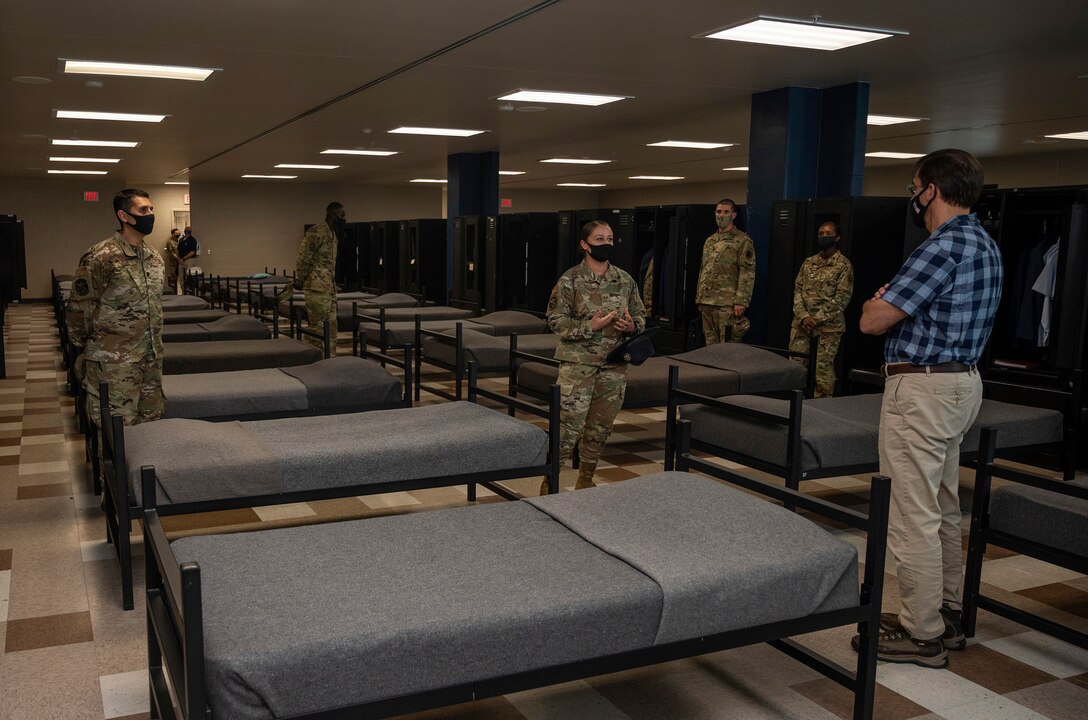 An airman briefs a civilian as basic trainees stand beside their bunks.