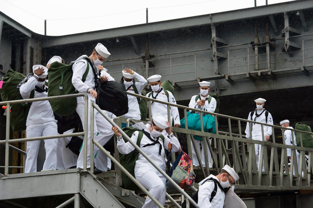 Sailors leave a ship wearing protective gear and carrying luggage.