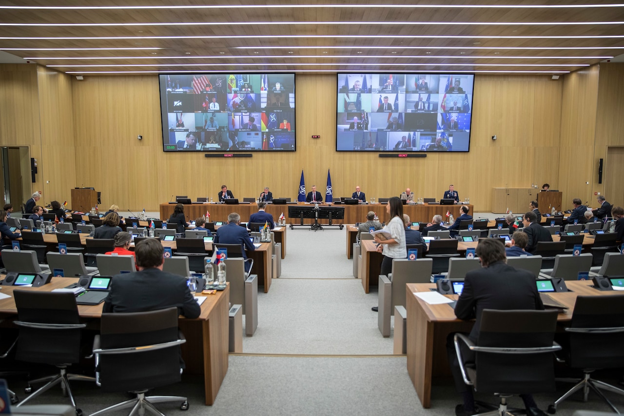 Screens display the faces of participants from around the world during a meeting as other participants practice social distancing in a large auditorium.
