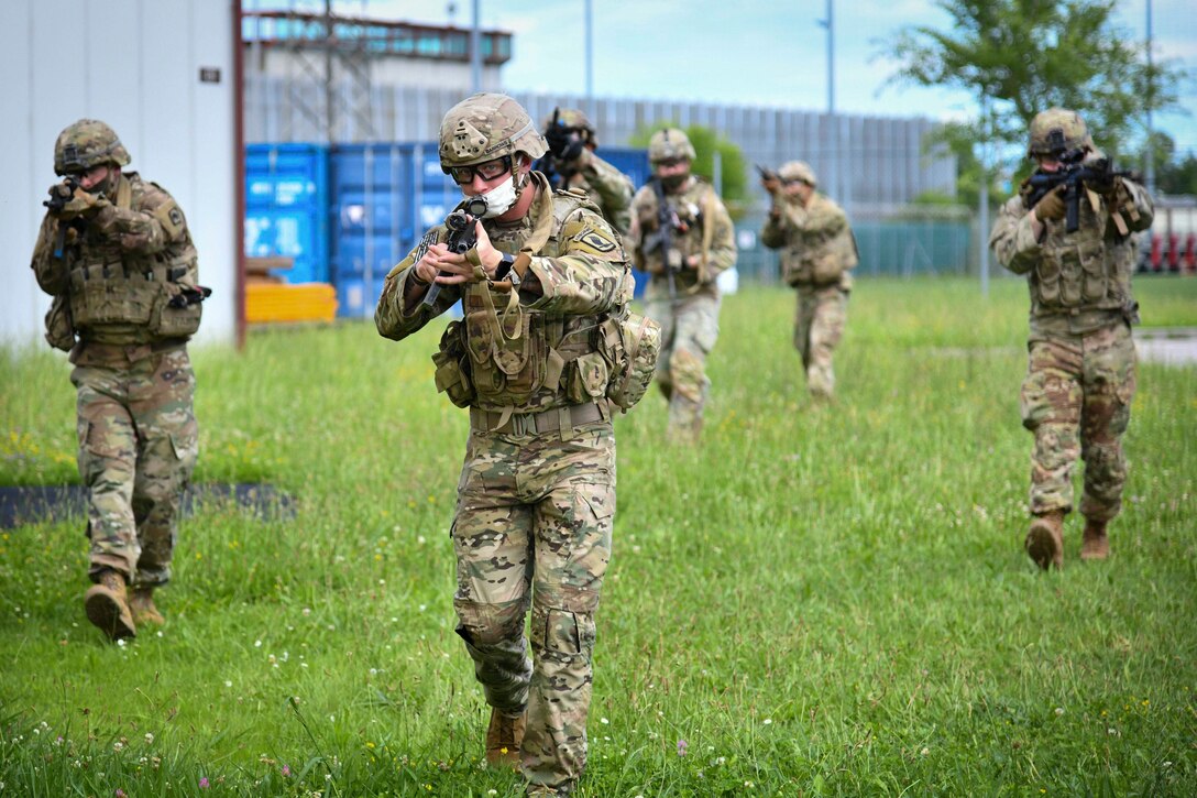 Soldiers wearing face masks walk through grass holding up weapons.
