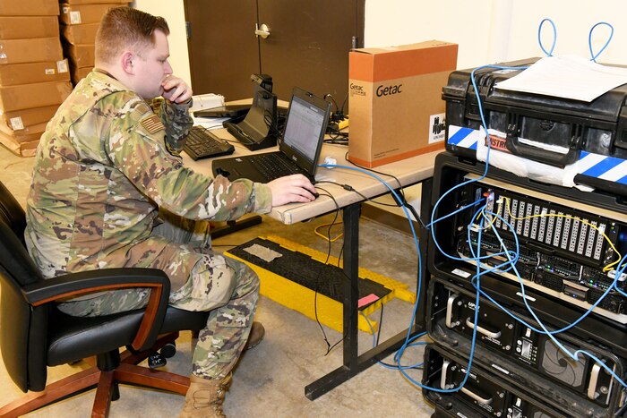 Photo shows an Airman working on a laptop connected to a tower of communications equipment.