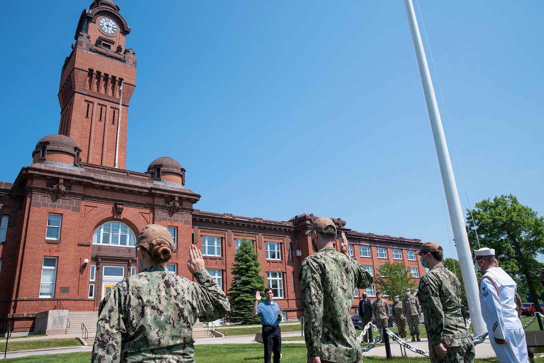 Defense Secretary Dr. Mark T. Esper stands with his right hand raised outside a brick building in front of sailors with theirs raised too.