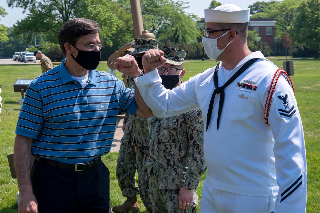 Defense Secretary Dr. Mark T. Esper bumps elbows with a sailor while wearing a mask and standing outside with other troops.