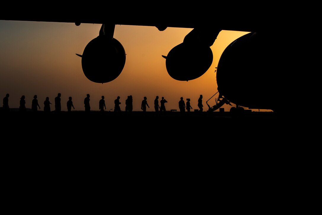 A line of service members are silhouetted against an orange sky as they prepare to board a large military aircraft.