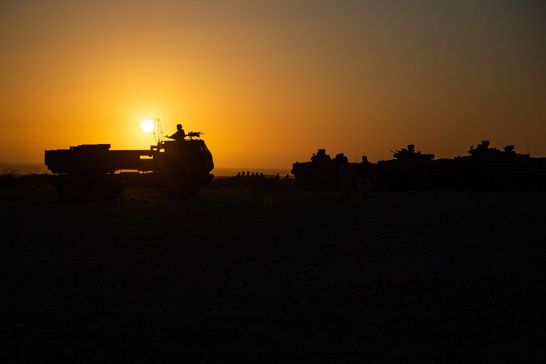 A service member, shown in silhouette, sits in the turret of a large military vehicle, illuminated by orange sky.