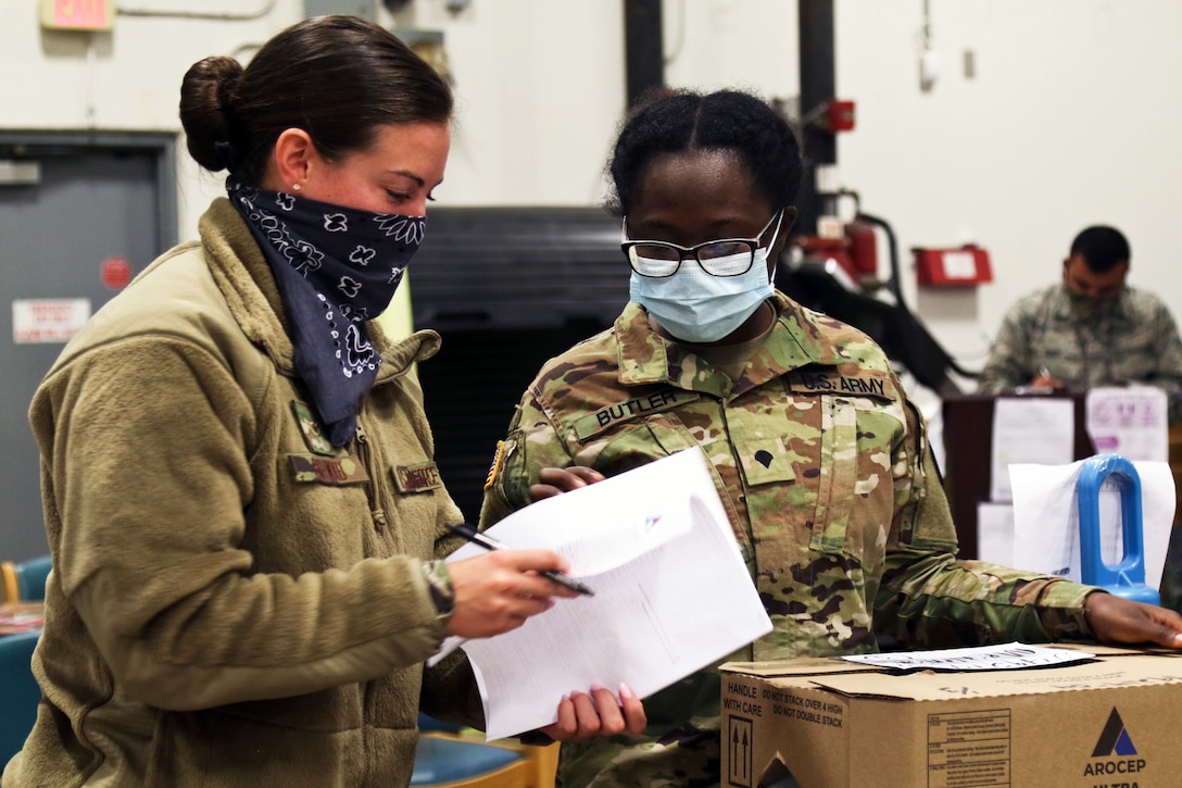 Two service members wearing face masks stand in a warehouse and look at some papers.