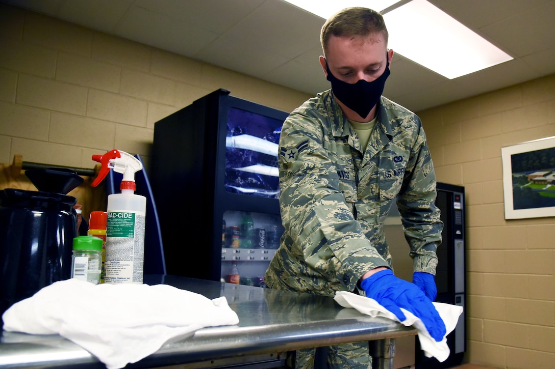 A service member, who is wearing a face mask and gloves, wipes a tabletop.