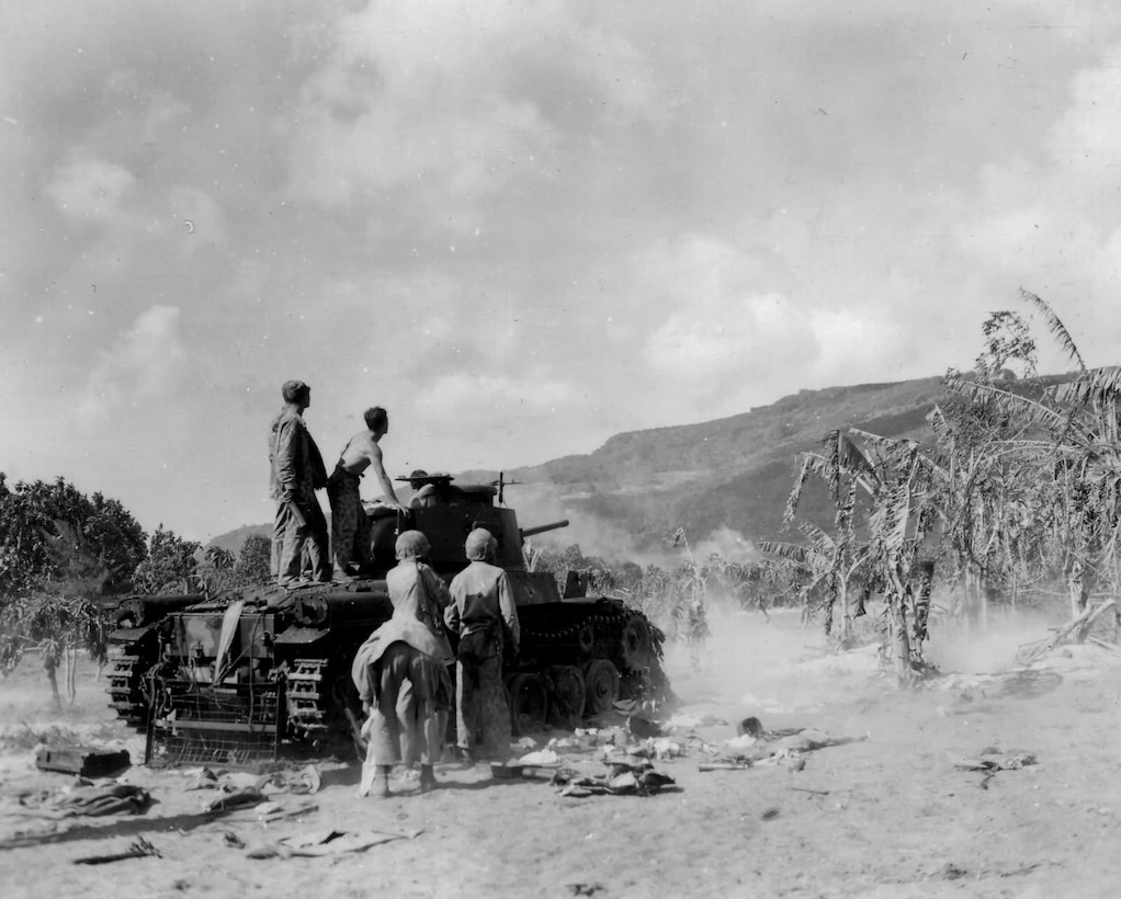 Five men stand on and around a captured Japanese World War II tank near palm trees and a mountain.