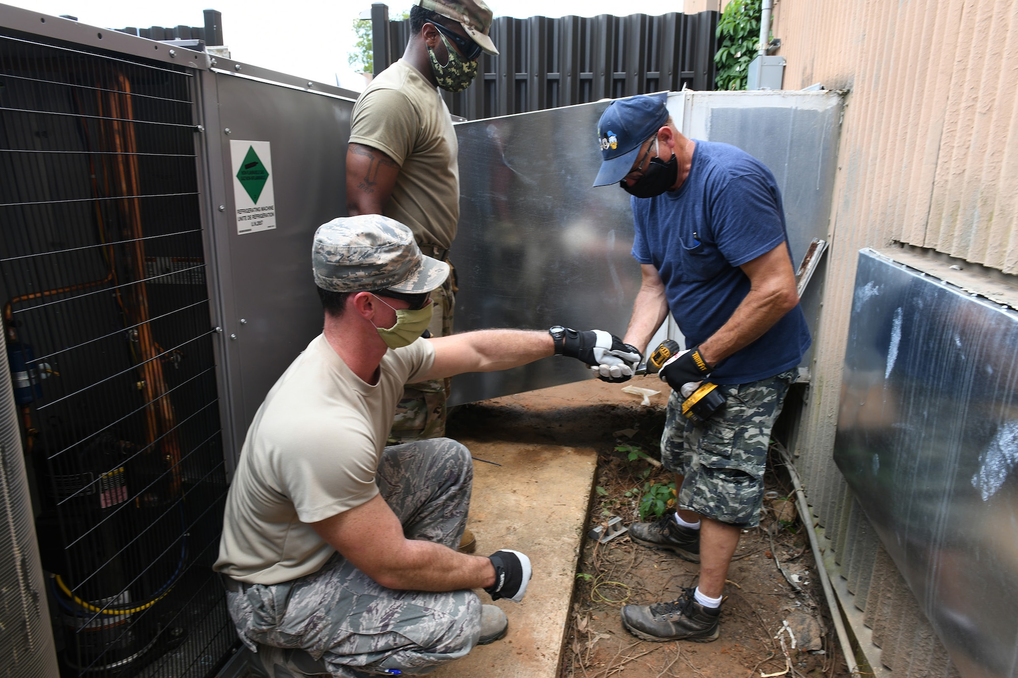 Photo shows two Airmen and a civilian working together on a HVAC unit outside.