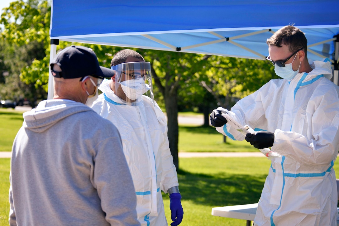 Two people in white coveralls and other protective gear talk to a civilian wearing a mask.