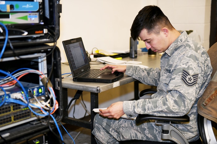 Photo shows an Airman working on a laptop next to a stack of boxes with wires coming out.