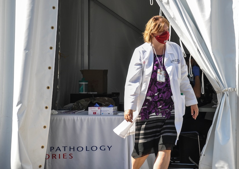 A woman wearing a face mask and a white lab coat walks out of a tent.
