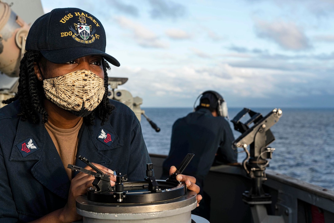 A sailor wearing a face mask stands with her hands on a round metal instrument and looks out on a ship's deck.