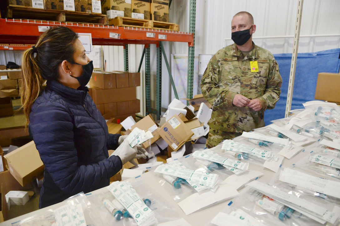 Two National Guardsmen wearing face masks stand next to a table of medical test kits.