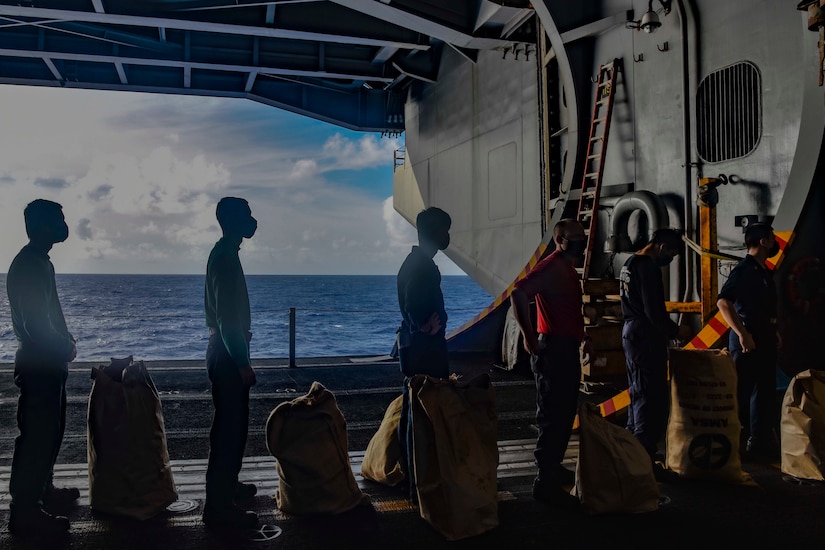 Sailors line up inside a ship. Each has a large bag sitting in front of them.
