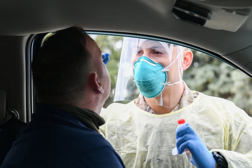 A man wearing a blue mask tests a motorist for COVID-19 through the driver’s window.