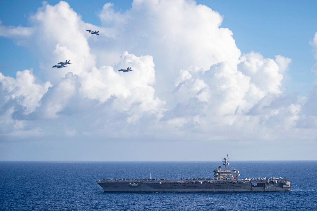 Four jets fly above an aircraft carrier traveling in blue seas.