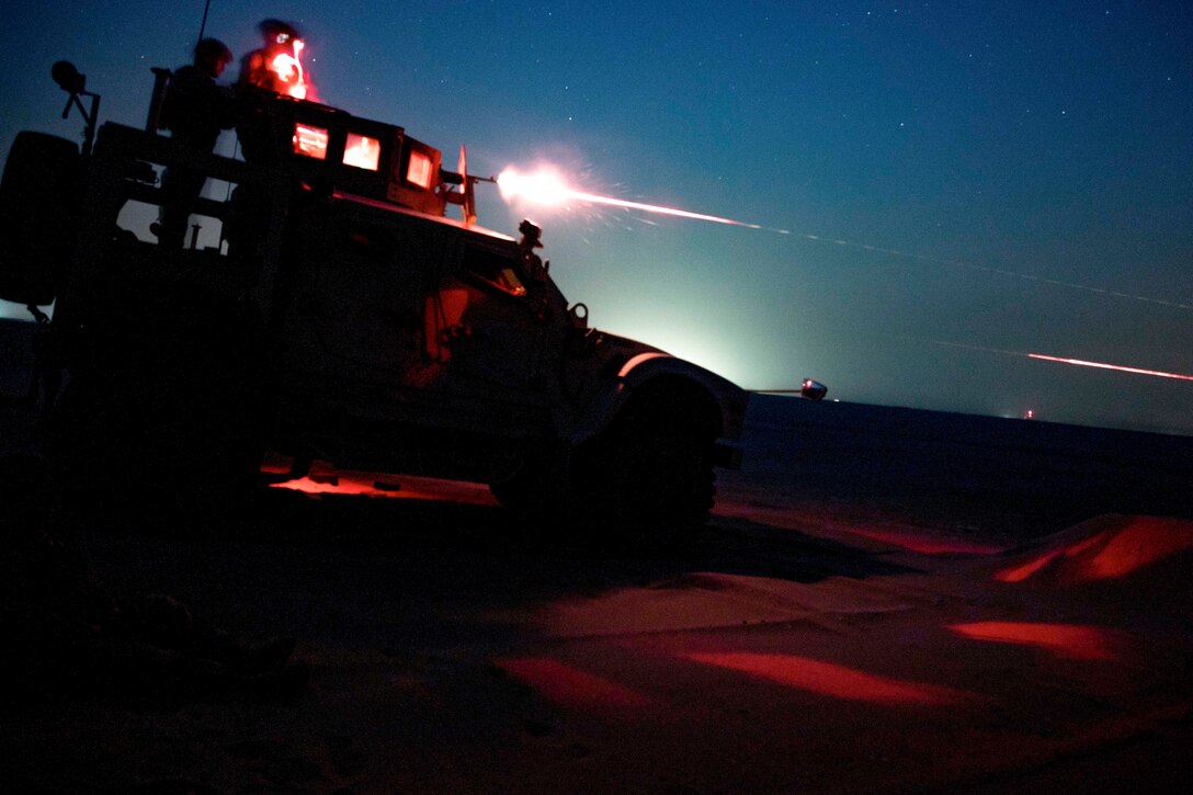 Marines, shown in silhouette and illuminated by red light, stand atop a military vehicle and fire a weapon at night.