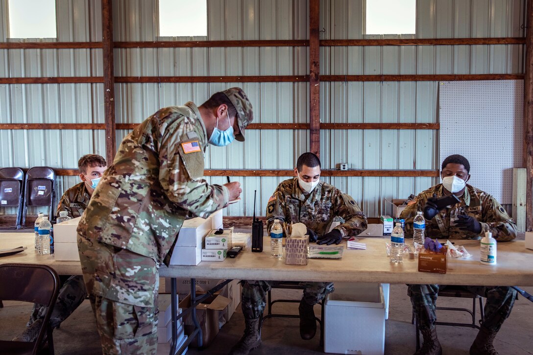 Service members wearing masks sit at a long table as a service member in the foreground writes on a pad.