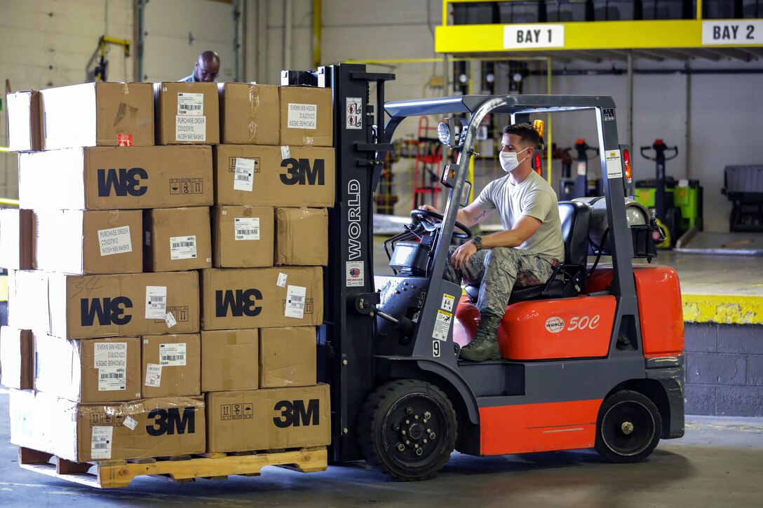 A service member wearing a mask moves boxes with a forklift as a man in the background looks on.with a fork-lift.