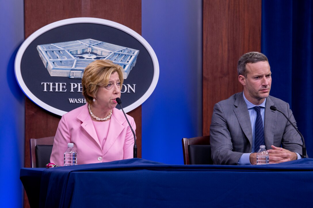 A woman speaks to reporters while a man sits next to her.