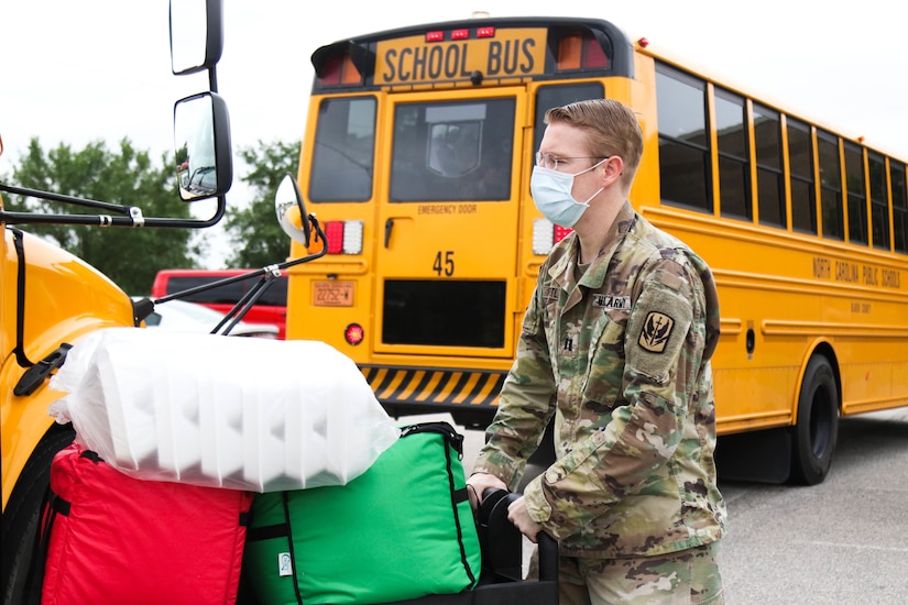 A soldier wearing a face mask pushes a cart loaded with packaged meals.