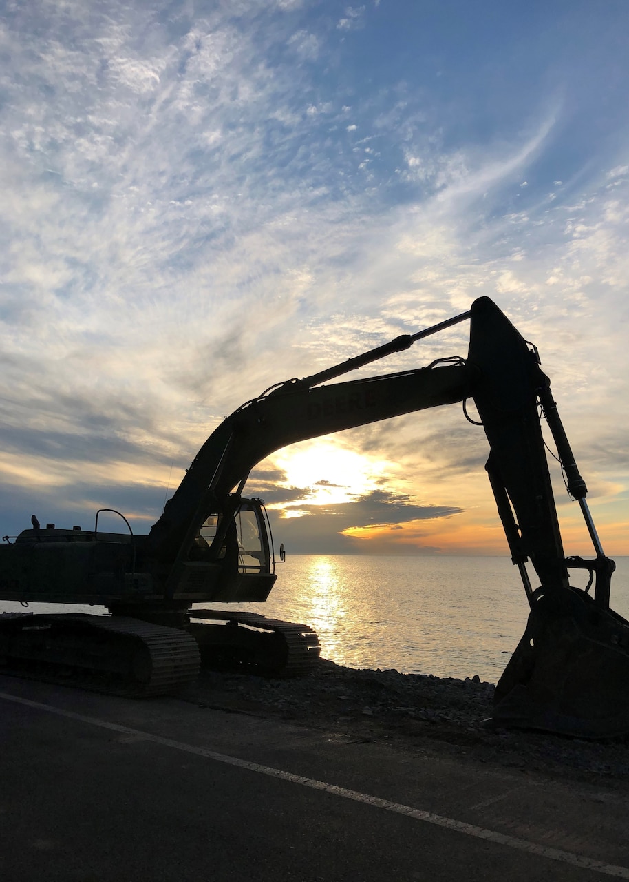 A steam shovel operates, silhouetted by a low sun over water in the background.