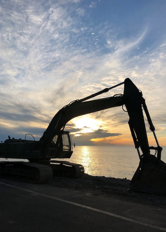 A steam shovel operates, silhouetted by a low sun over water in the background.