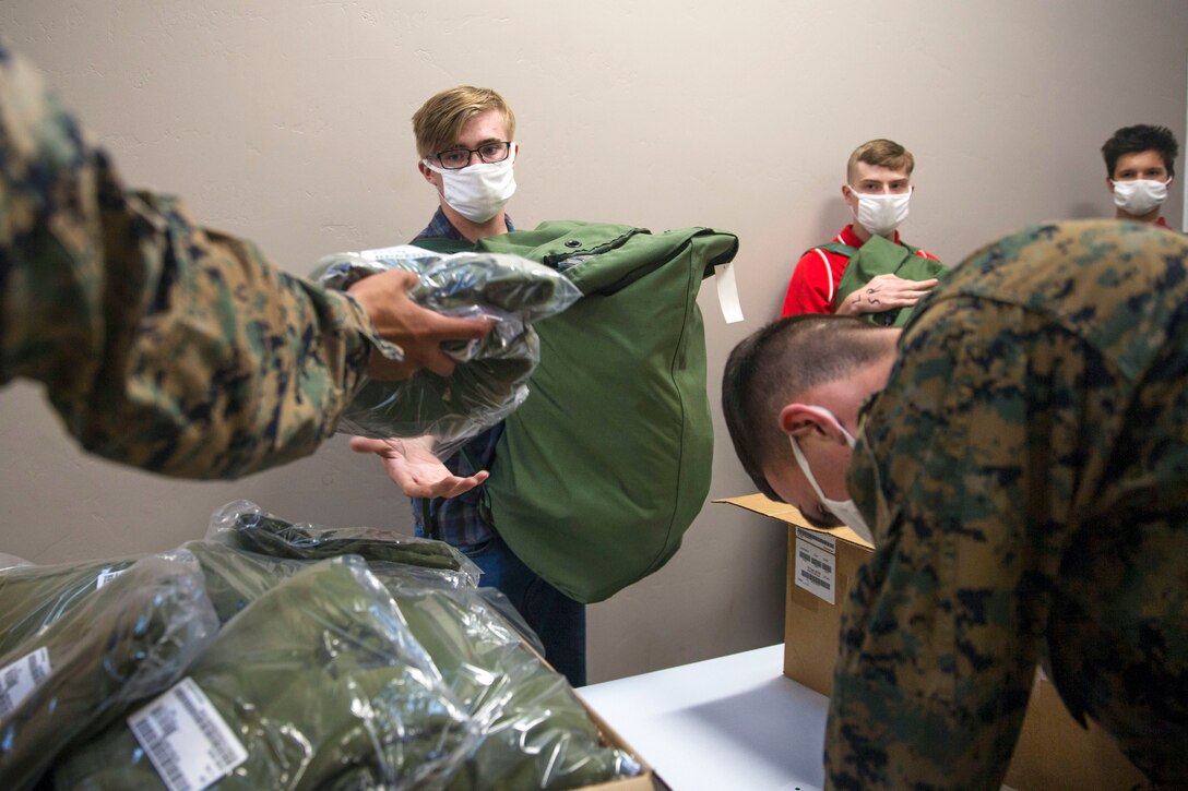 A Marine recruit wearing a mask reaches for gear while other recruits wearing masks stand socially distanced waiting their turn.