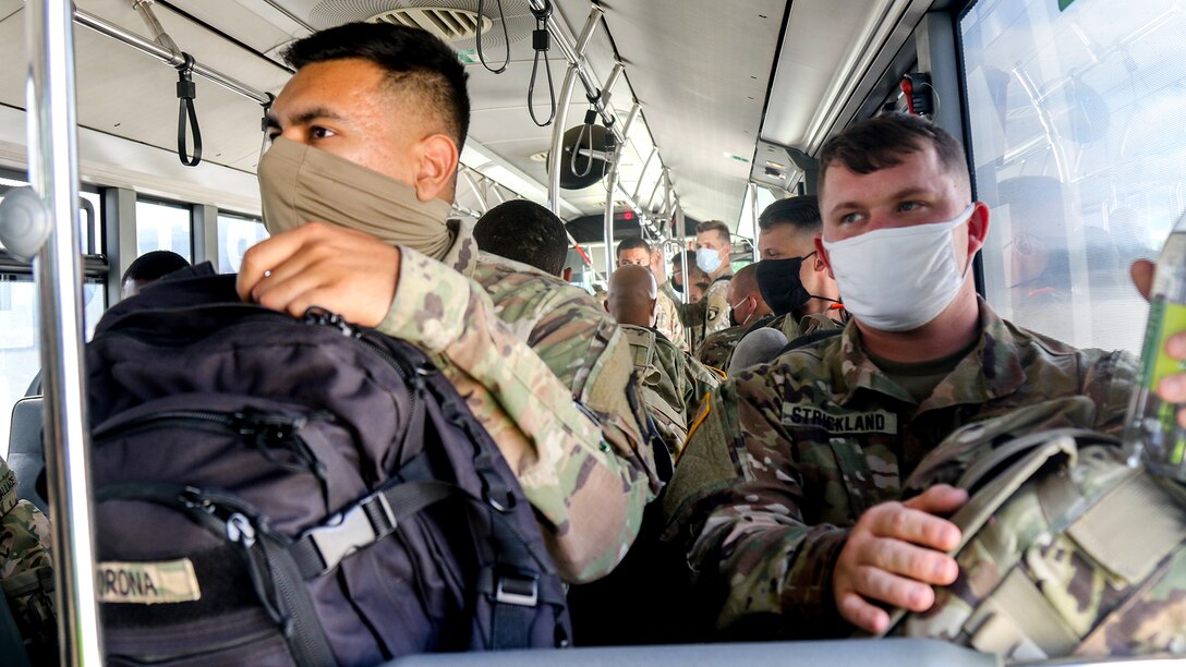 Two soldiers wearing face masks and holding backpacks on their laps sit in a bus.