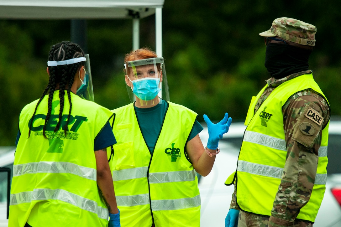 Standing outside with a car in the background, a soldier wearing personal protective equipment speaks to two other people wearing face shields and gloves.