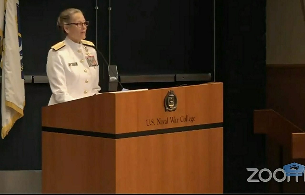 A woman wearing a Navy uniform stands at a lectern and speaks.