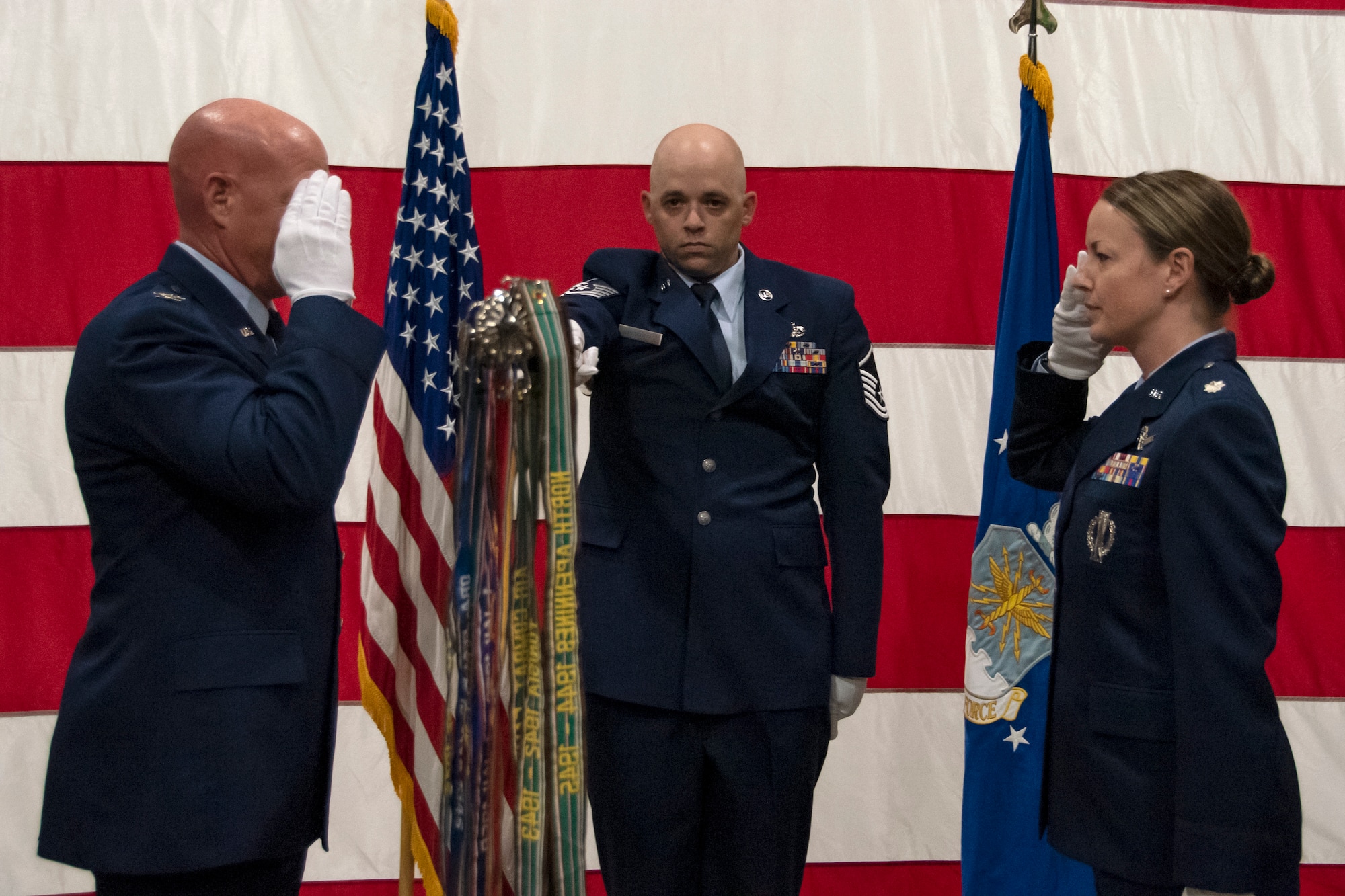 A man and woman salute each other while a third man extends out a flag straight out at should-length.