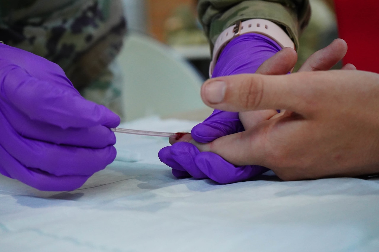 A soldier wearing latex gloves takes a sample of blood from someone’s finger.