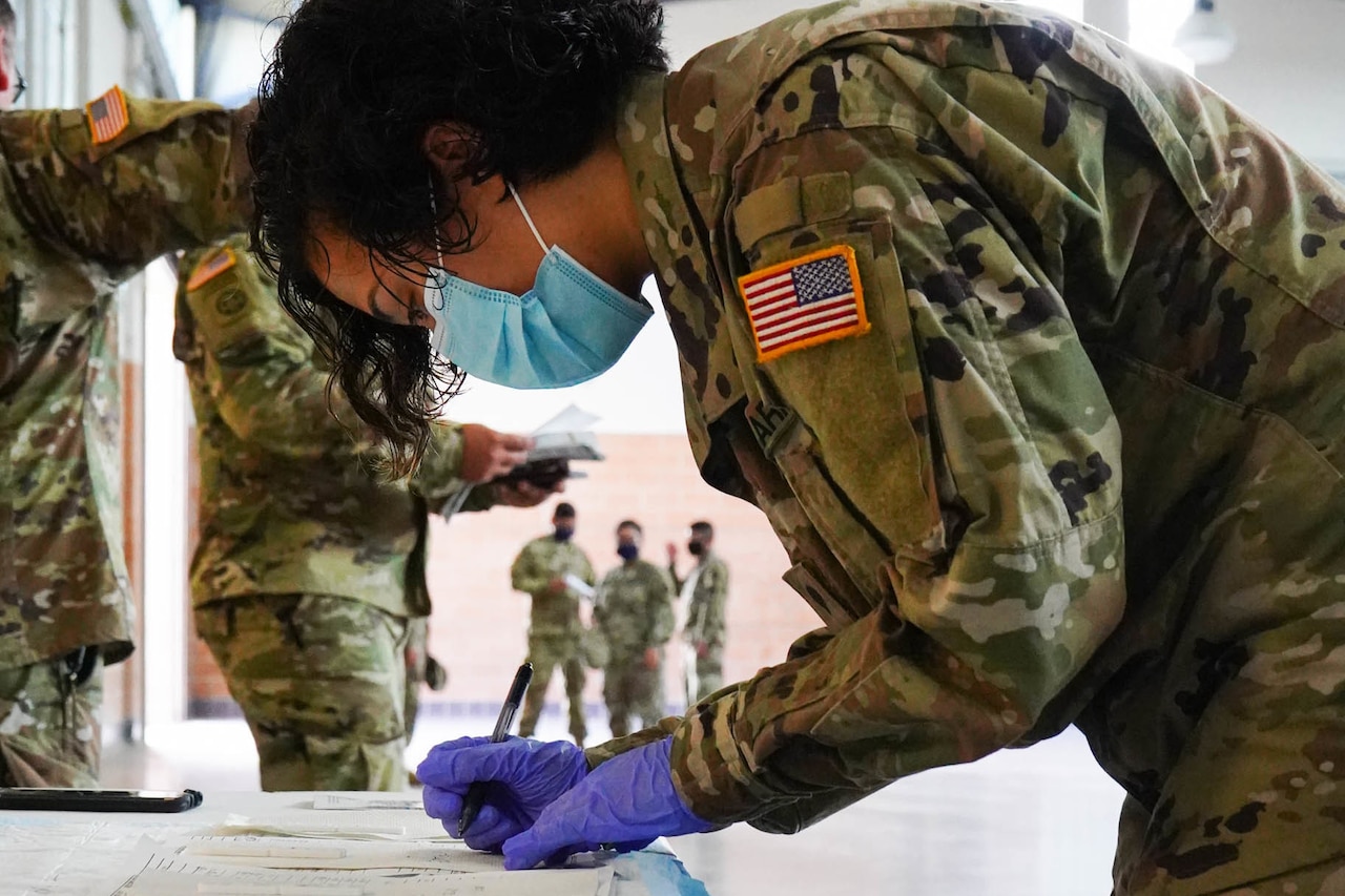 A soldier wearing a face mask and latex gloves leans at a table to write on a form.