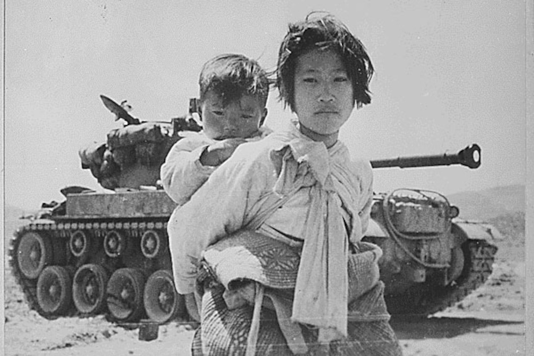 Young girl in traditional Korean clothes carries her baby brother. A tank is in the background.