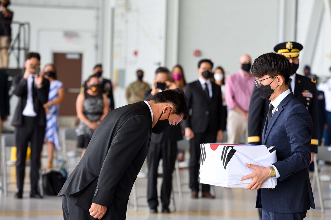 A South Korean official bows at the waist as another official holds a box wrapped in a South Korea flag.
