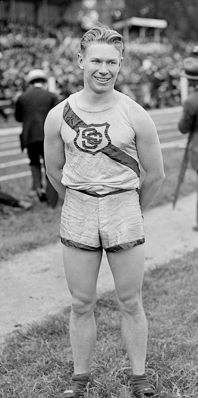 A young man wearing shorts and a T-shirt emblazoned with a team logo poses for a photo standing outside on an athletic field.