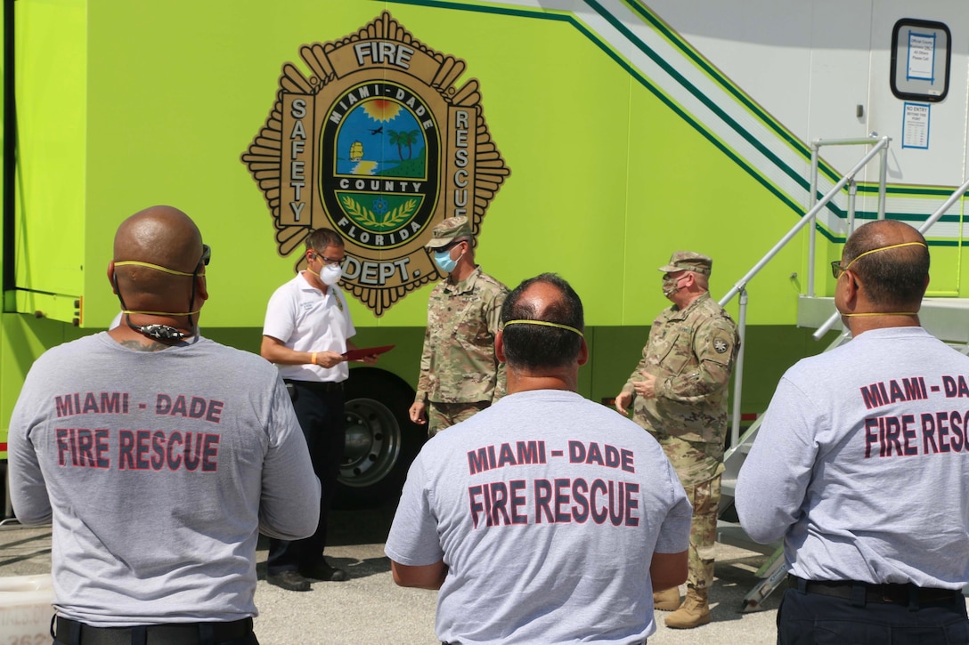 Two soldiers congratulate a civilian as men wearing Miami-Dade County Fire Rescue T-shirts look on.