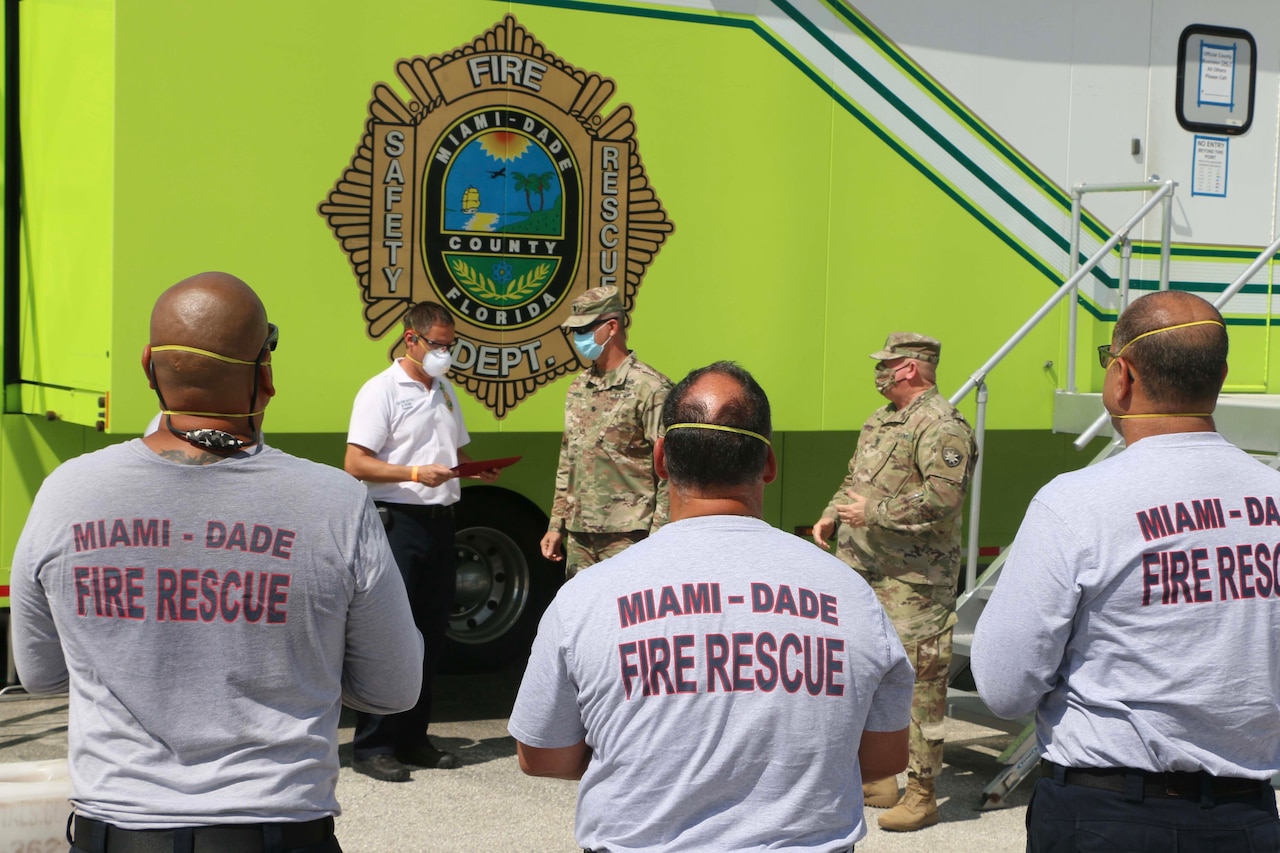 Two soldiers congratulate a civilian as men wearing Miami-Dade County Fire Rescue T-shirts look on.