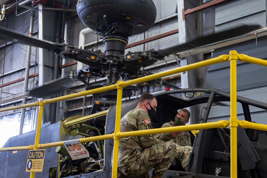 Sergeant Major of the Army sits in an AH-64 Apache helicopter trainer as a Soldier briefs him on the equipment.