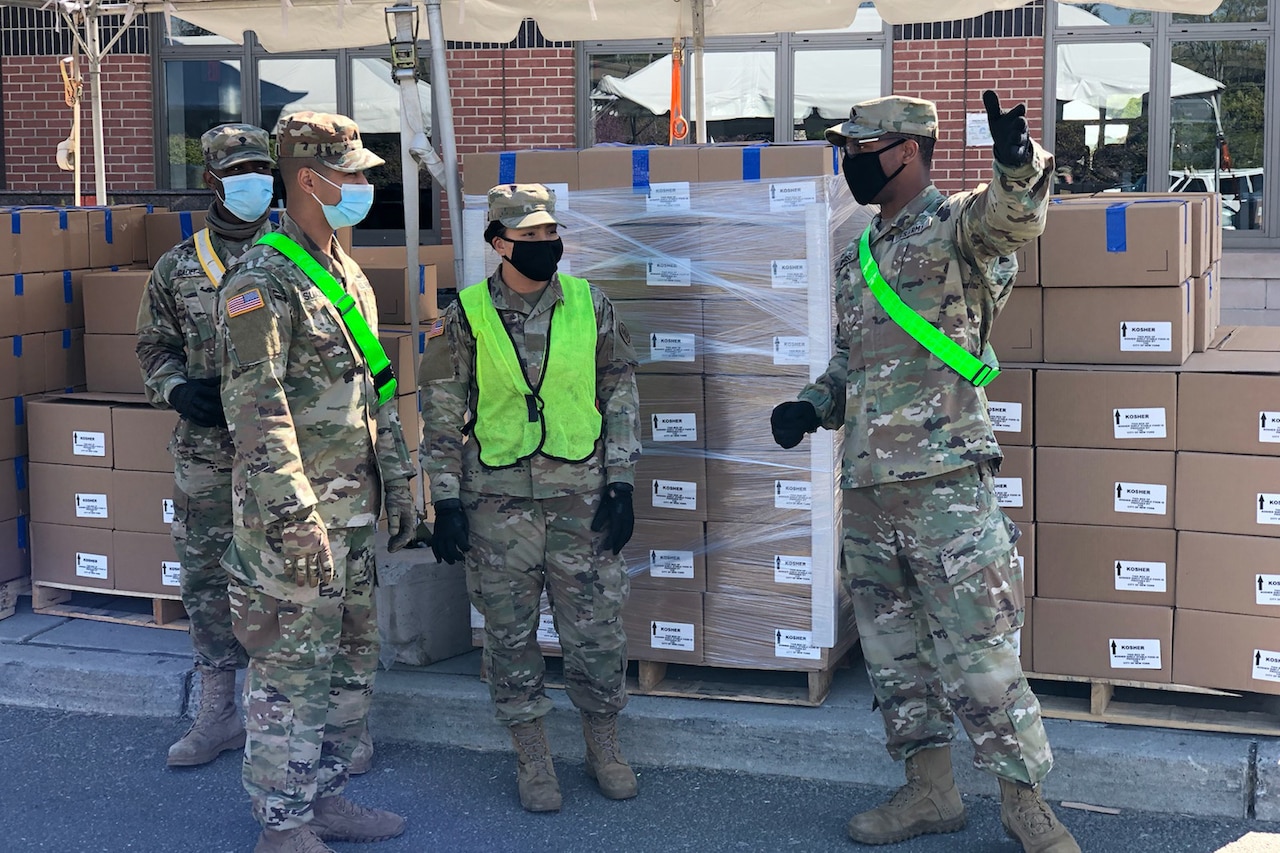 A soldier gestures as he gives guidance to other soldiers in front of stacks of cardboard boxes.