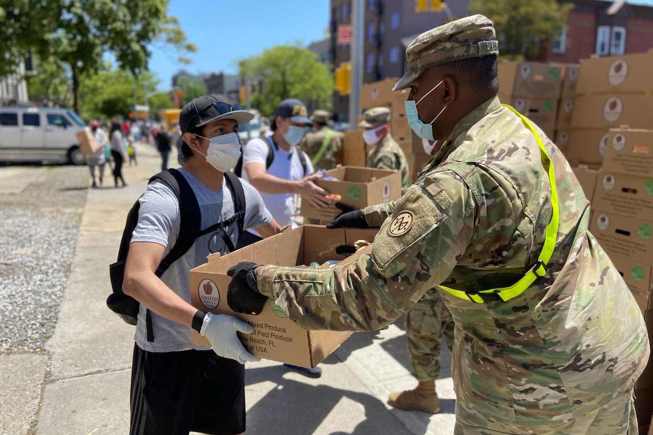 A soldier wearing a mask hands a box of food to a civilian in a mask.
