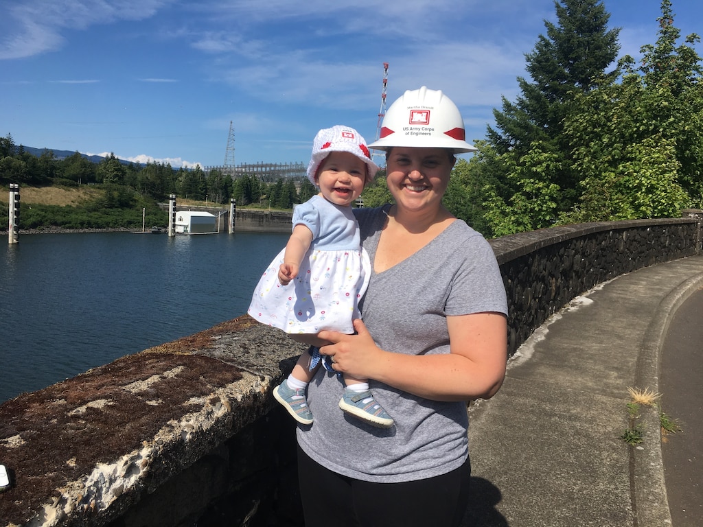 A woman in a hard hat holds a baby in a bonnet near Bonneville Dam.
