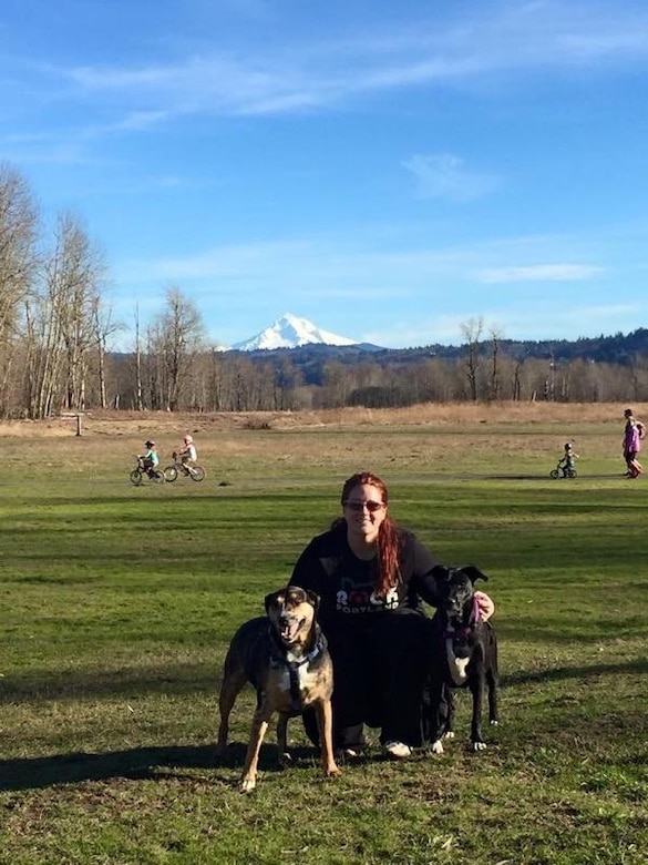 A woman poses in a grassy field with two large dogs. A snowy mountain is in the background.