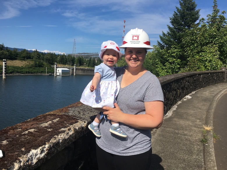 A woman in a hard hat holds a baby in a bonnet near Bonneville Dam.