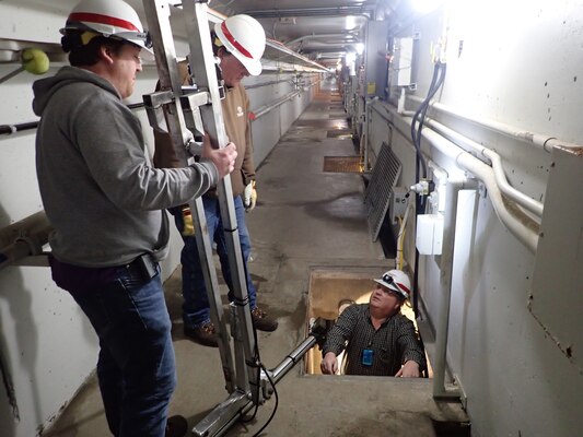 2 men stand in long corridor with strange device, overlooking third man in a rectangular hole in the floor