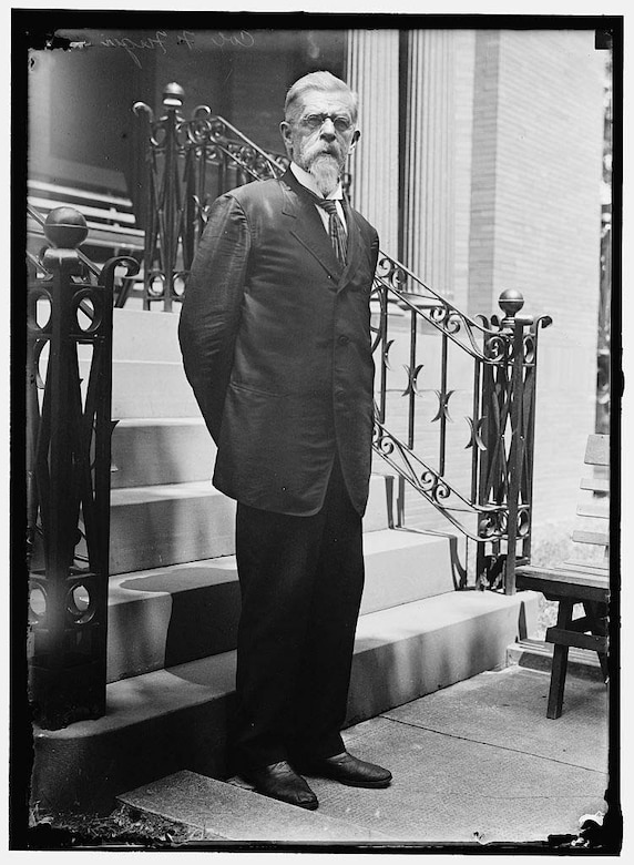 An older man stands with his hands behind his back at the foot of six exterior stairs.